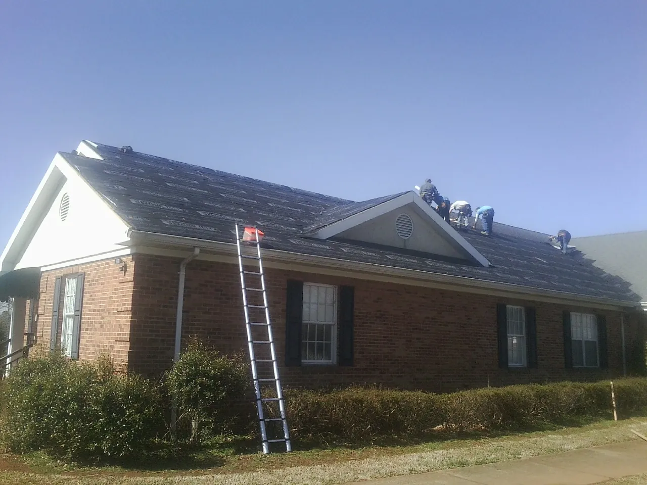 Crew tearing shingles off a brick ranch roof with a ladder set — Emergency Roof Repair work by our crew in Henry
