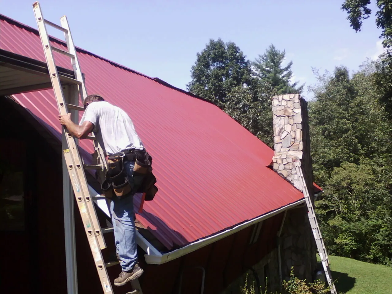 Roofer on a ladder beside a red metal roof and stone chimney — Soffit & Fascia Repair by Roofing Brothers in Henry